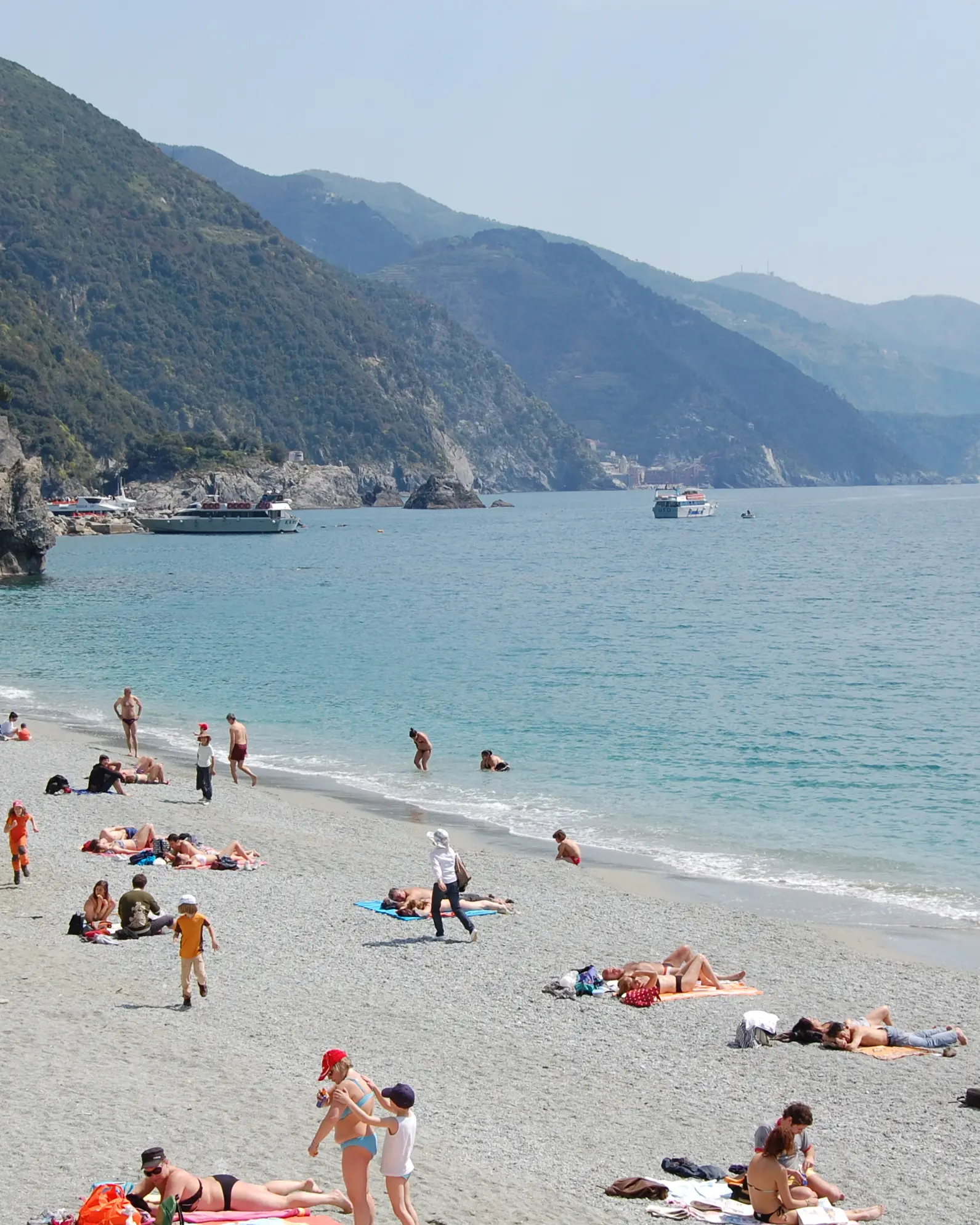 Beachgoers beside turquoise water with boats and coastal mountains in the distance